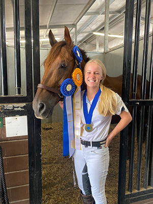 A girl standing next to her horse, smiling at the camera. The horse has many ribbons surrounding it.