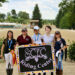 Five girls holding a sign standing in front of the camera, smiling.