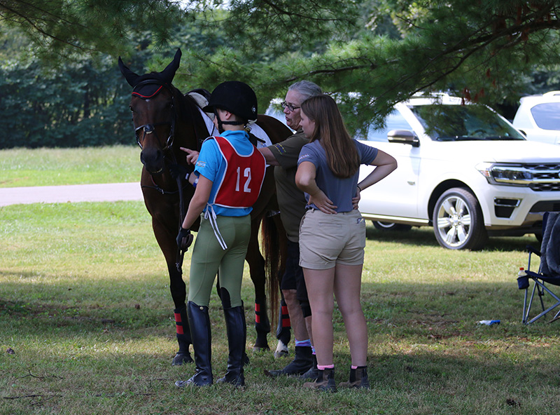 A group of three people standing at a horse, one is pointing out something on the horse's tack.