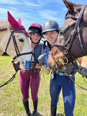 Two individuals and their horses, smiling at the camera and holding blue ribbons.