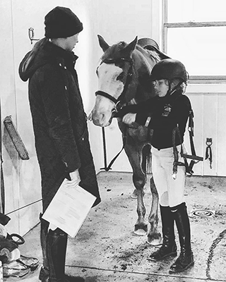 A young girl tacking up her horse with her instructor watching.