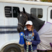 A young girl and her horse standing at a horse trailer, holding a blue ribbon and smiling at the camera.