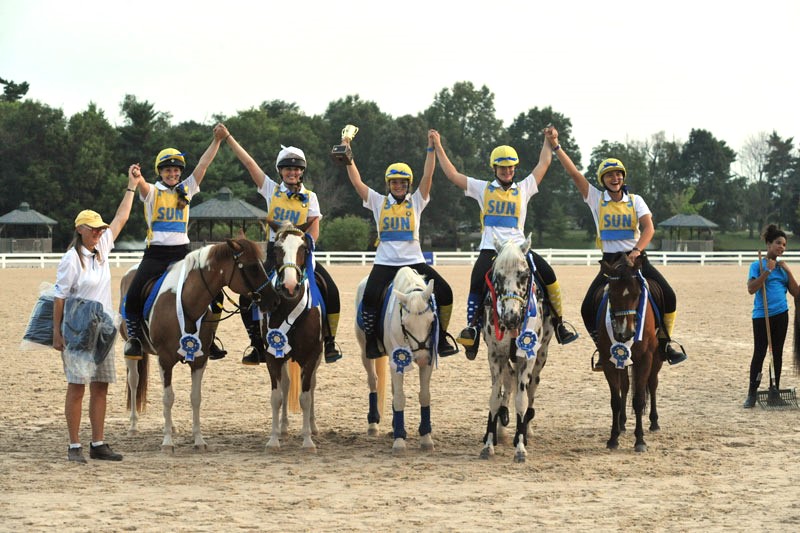 team of five riders holding hands up in the air while mounted on ponies wearing blue ribbons