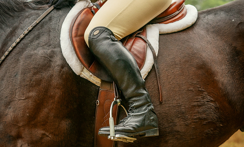 A person riding a horse, the image is zoomed in to just the saddle and the left leg of the rider.
