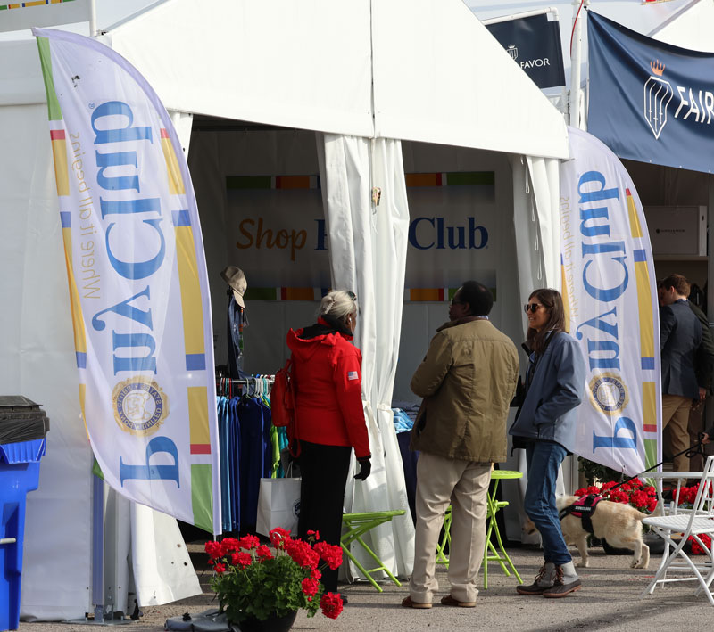 visitors outside the Pony Club trade fair booth in a white tent with Pony Club banners and red geraniums in pots