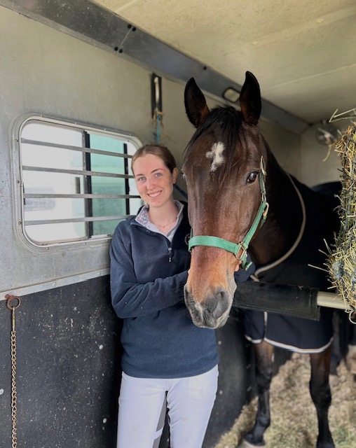 A young woman in a horse trailer, smiling for the camera with her horse.